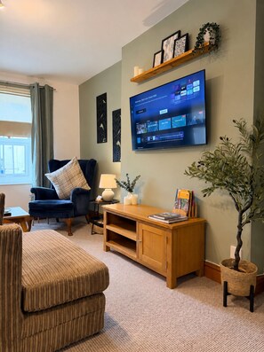 A living room with a television mounted on the wall, a blue armchair, a wooden coffee table, and a bookshelf. - Solway Retreat, Cosy 2 Bed Flat, Sleeps 4 - M6 Jct45, A75, Gretna & Carlisle (Longtown)