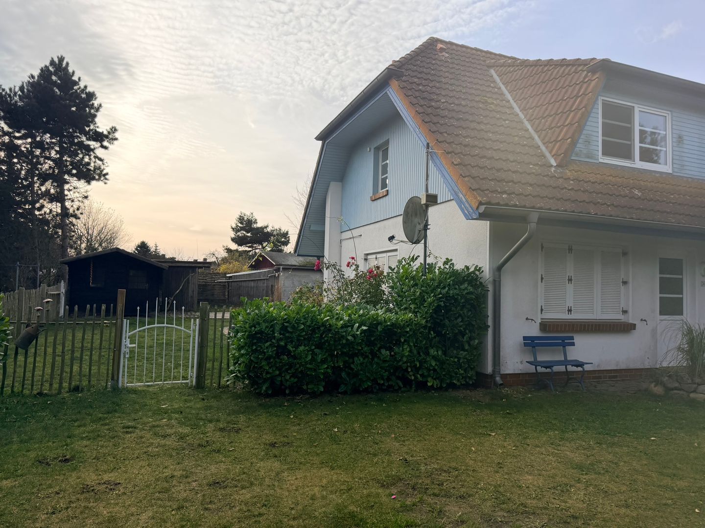 Una casa residencial con techo inclinado, antena parabólica y un pequeño jardín con un banco.