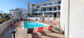 Un espace piscine avec des chaises longues et des parasols, un escalier menant à un bâtiment, et un ciel bleu dégagé.