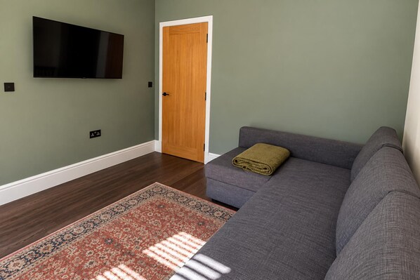 A living room with a grey sofa, a wooden door, a flat-screen TV mounted on the wall, and a patterned rug on the floor.