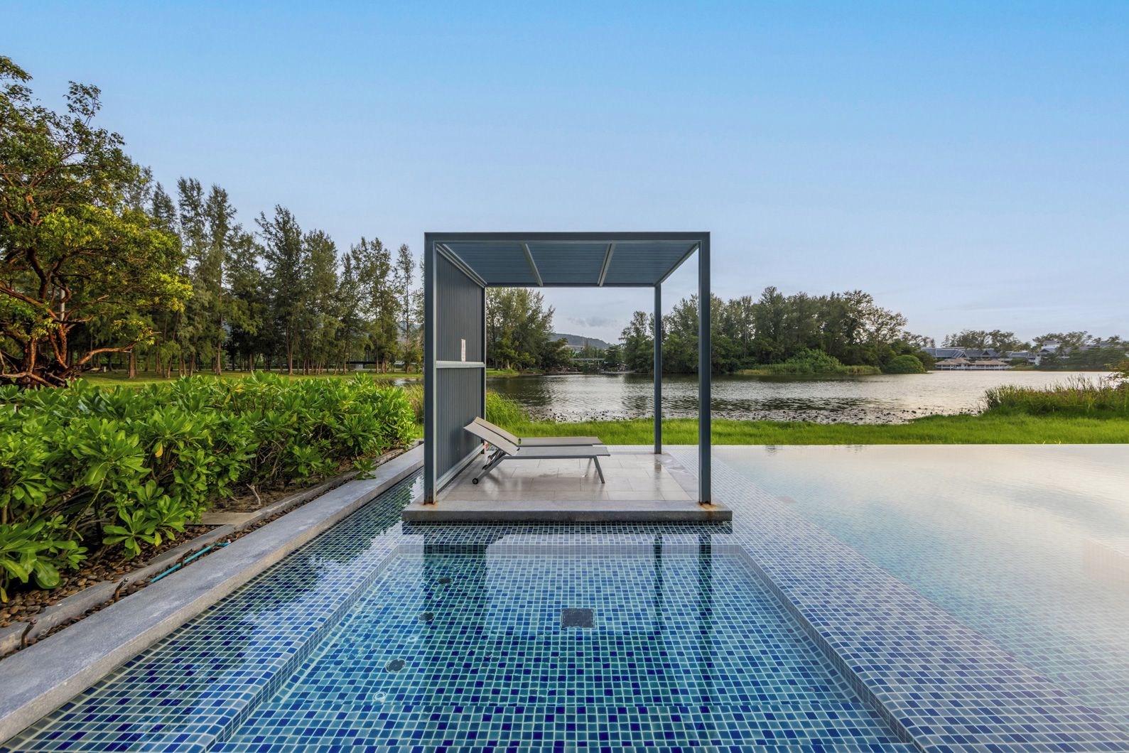 A modern outdoor pool area with a glass enclosure and a bench, surrounded by greenery and a view of a lake.