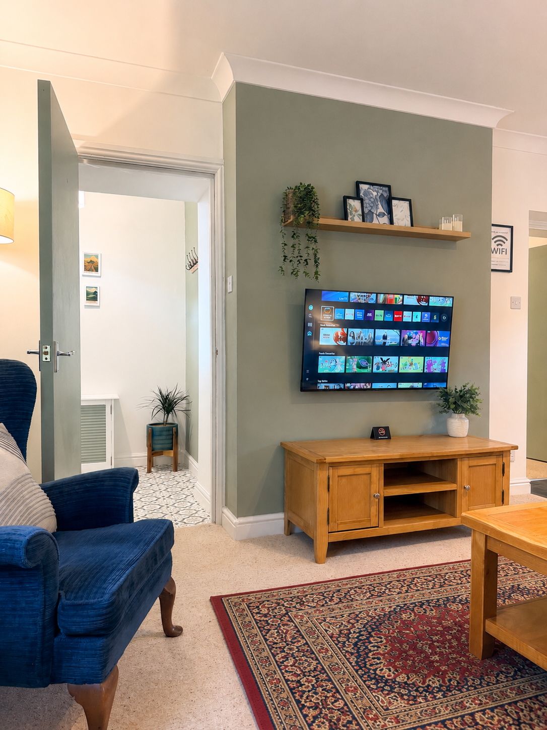 A living room with a flat-screen TV mounted on the wall, a wooden entertainment unit, a blue armchair, and a patterned rug.