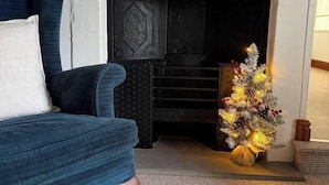 A living room with a blue armchair, a fireplace with a decorative mantle, and a small Christmas tree.