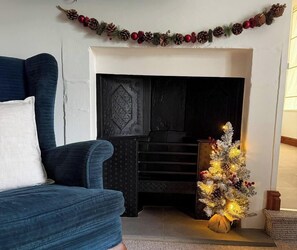 A living room with a blue armchair, a fireplace with a decorative mantle, and a small Christmas tree.