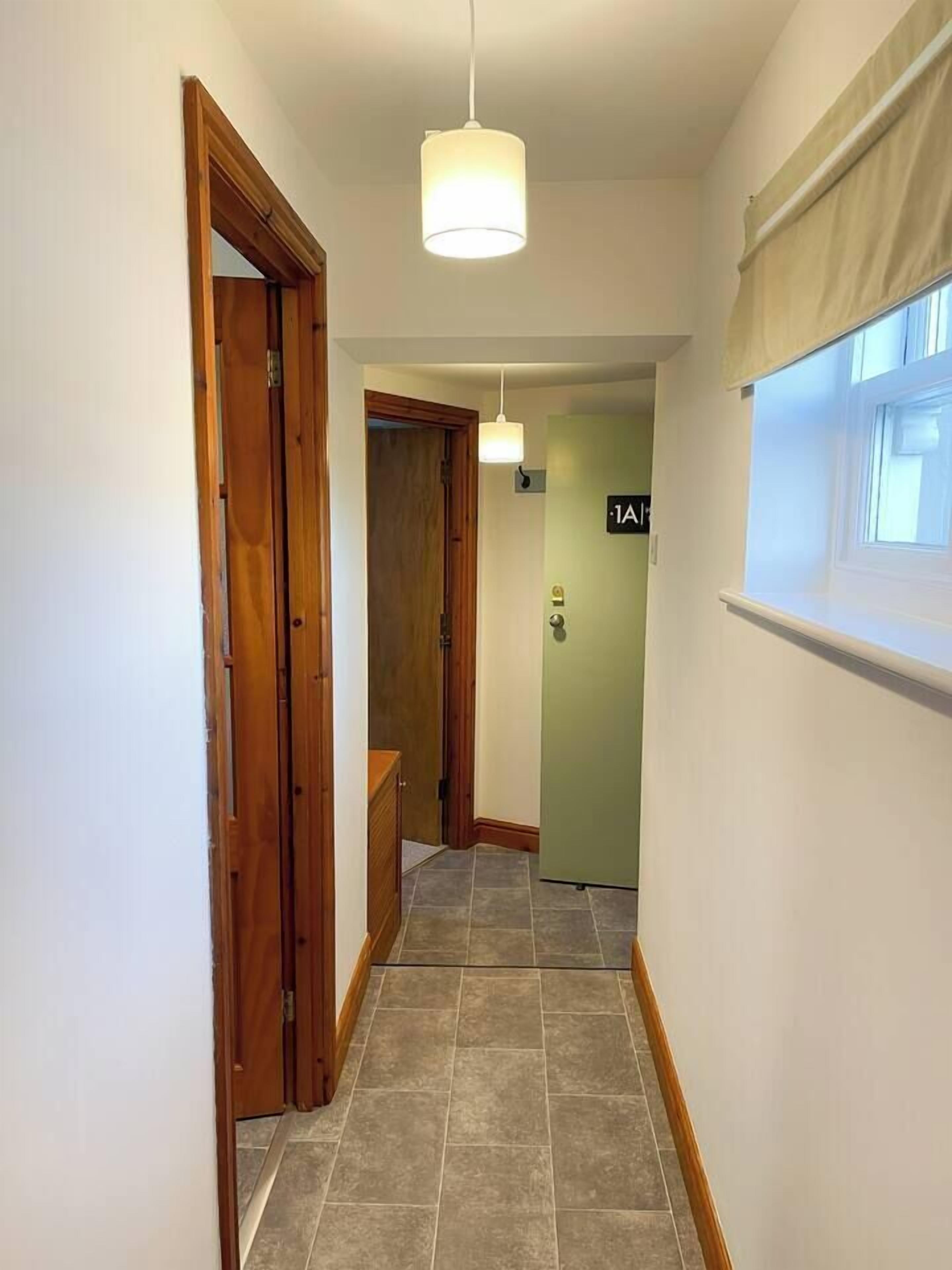 Un pasillo estrecho con puertas de madera, un aplique colgante y una ventana con persiana.