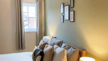 Ein Schlafzimmer mit einem Bett, einem Nachttisch mit Lampe, einem Fenster mit Vorhängen und gerahmten Bildern an der Wand.