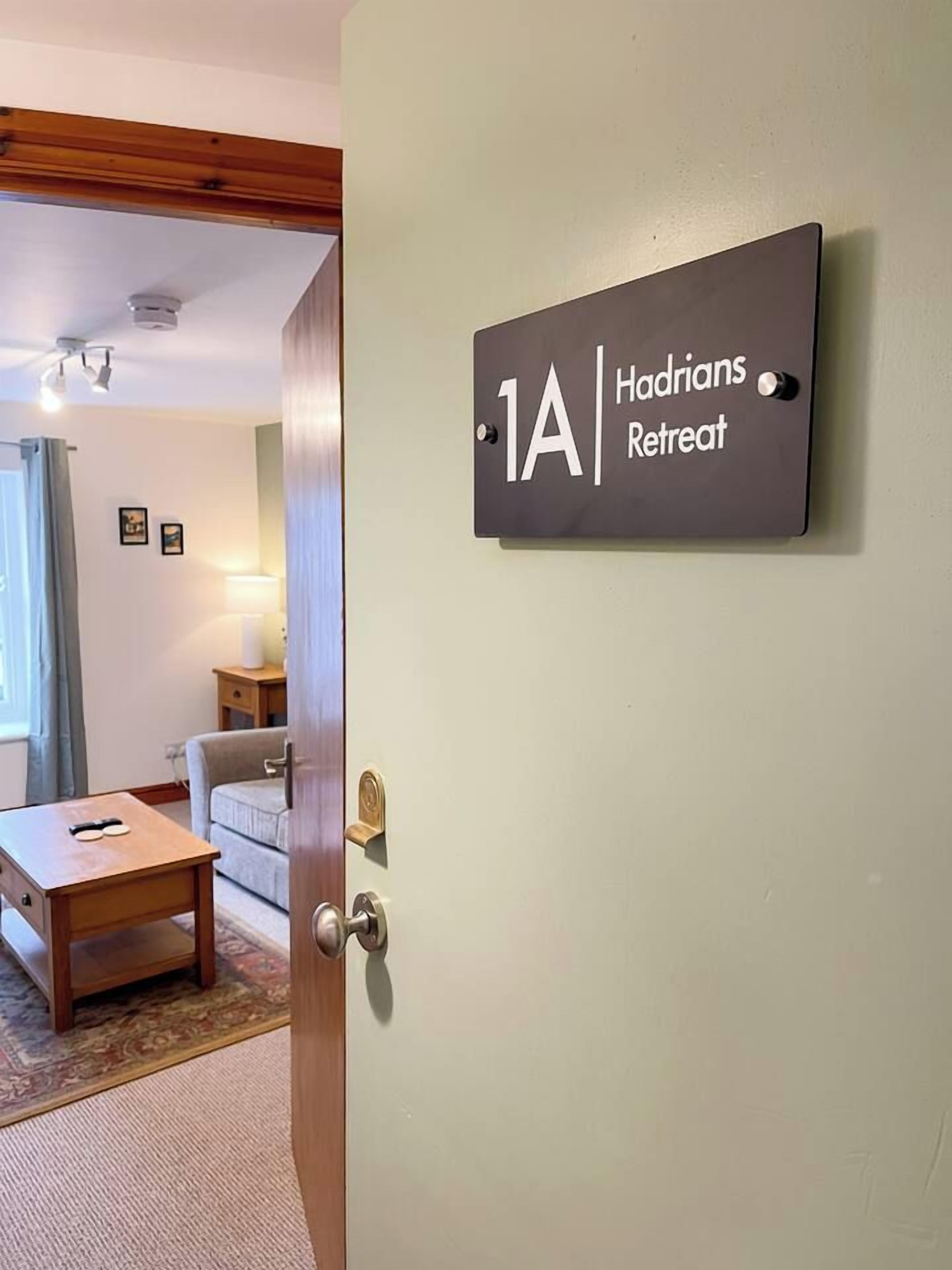 Una habitación con una puerta que indica '1A Hadrions Retreat' y parte visible de una sala de estar con una mesa y un sofá.