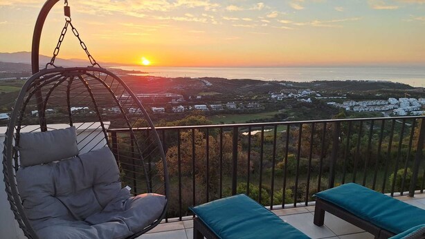 Een balkon met een hangstoel en twee lounge fauteuils, met uitzicht op een kustlandschap met gebouwen en heuvels.