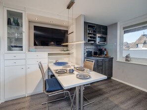 A modern kitchen with white cabinetry, a dining table set with blue and white plates, and a large flat-screen TV above the counter. - defaultName (Sylt)
