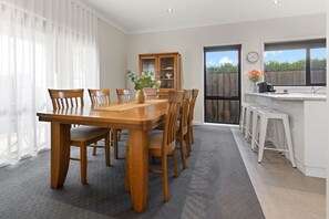 A dining area with a wooden table and chairs, a cabinet with glass doors, and a kitchen area with stools. - Avalon: A cosy, modern home that makes being away feel a little easier. (Bairnsdale)