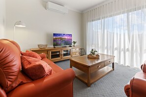 A modern living room with a leather sofa, wooden coffee table, and a flat-screen TV on a wooden cabinet. - Avalon: A cosy, modern home that makes being away feel a little easier. (Bairnsdale)