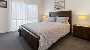 A bedroom with a large bed, two bedside tables, a headboard, a window with sheer curtains, and a painting on the wall.