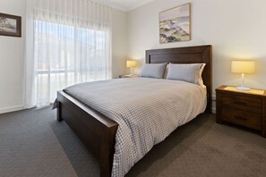 A bedroom with a large bed, two bedside tables, a headboard, a window with sheer curtains, and a painting on the wall. - Avalon: A cosy, modern home that makes being away feel a little easier. (Bairnsdale)