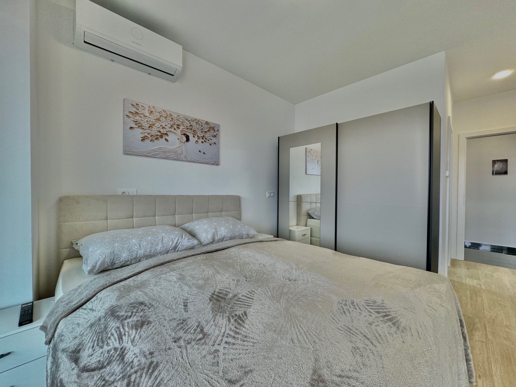 Une chambre à coucher moderne avec un lit, un climatiseur fixé au mur, un tableau représentant un arbre décoratif et une armoire.