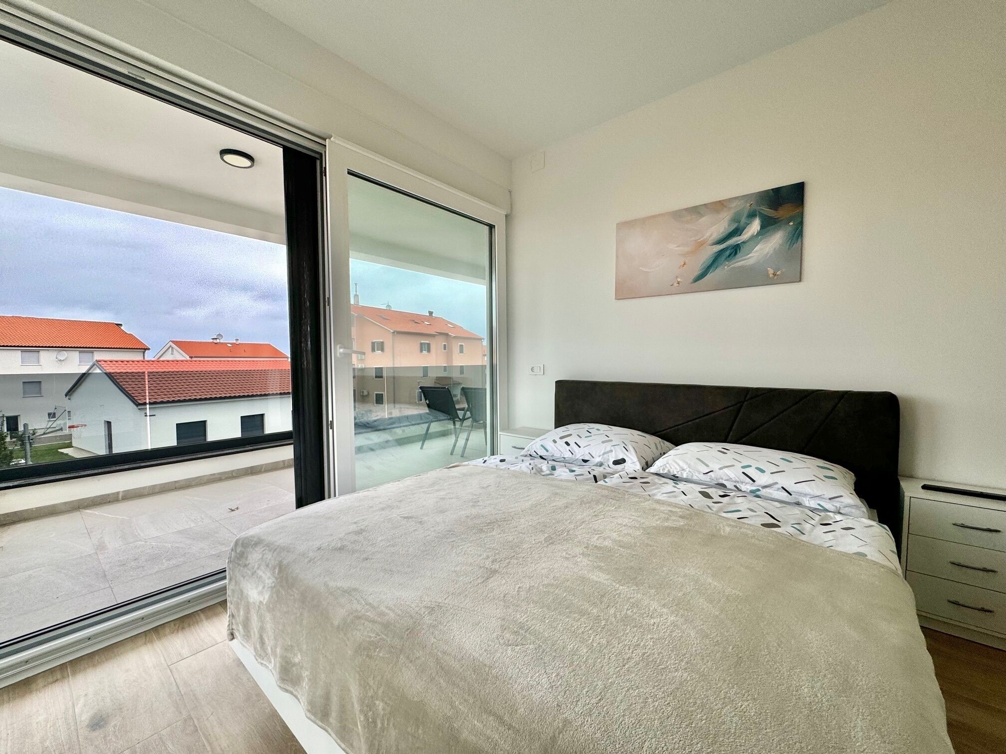 A bedroom with a large window, a bed with patterned bedding, a nightstand, and a wall-mounted artwork.