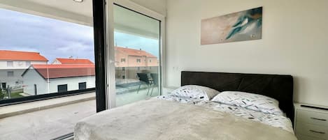 A bedroom with a large window, a bed with patterned bedding, a nightstand, and a wall-mounted artwork.
