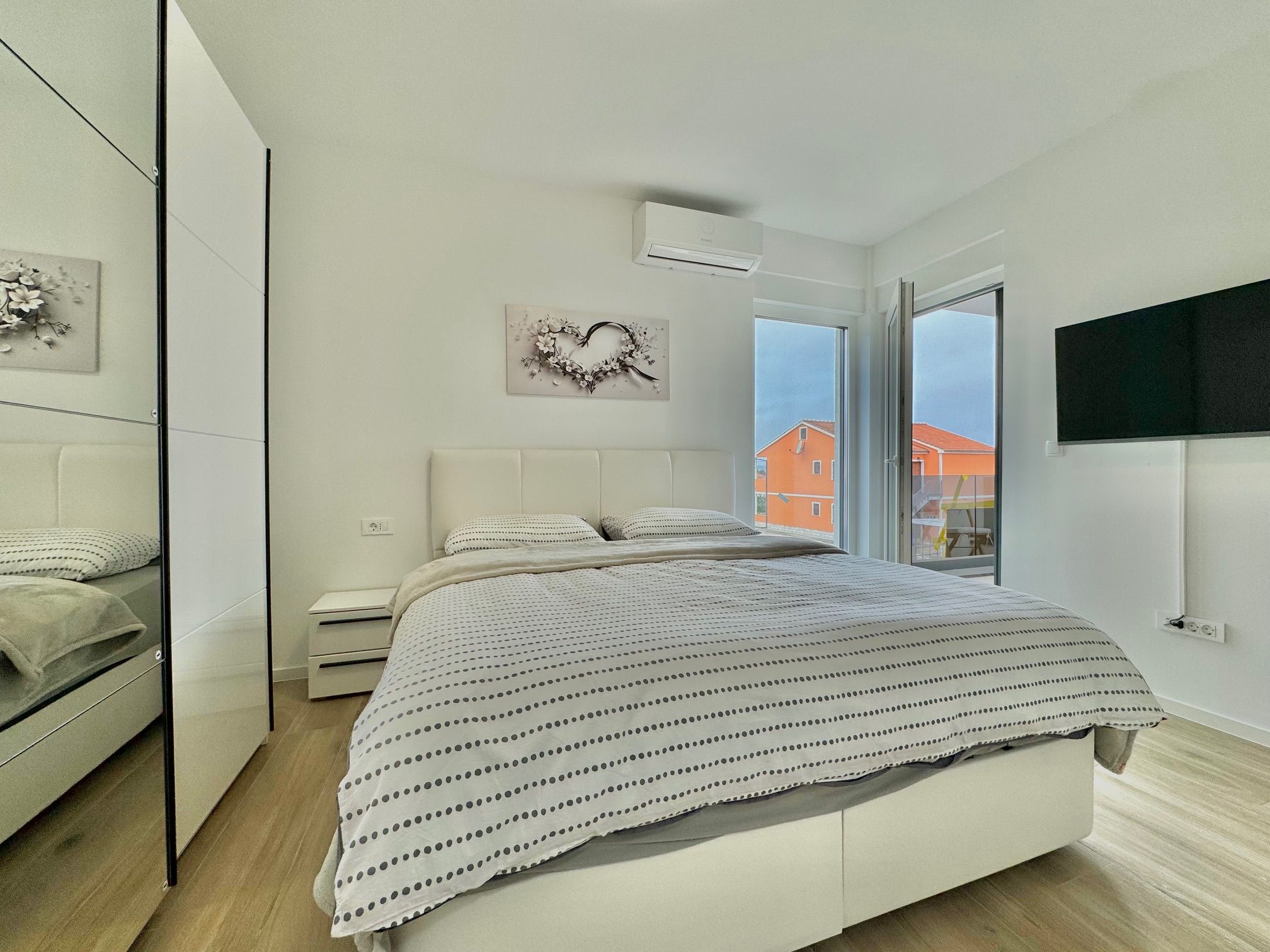 Een moderne slaapkamer met een bed, nachtkastjes, een dressoir, een wandtelevisie en een raam met uitzicht op gebouwen.