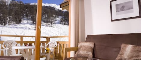 Une pièce chaleureuse offrant une vue sur un paysage montagneux enneigé. Elle comprend un canapé marron, une table à manger avec des éléments pour le petit-déjeuner et un balcon doté d'une balustrade en bois.