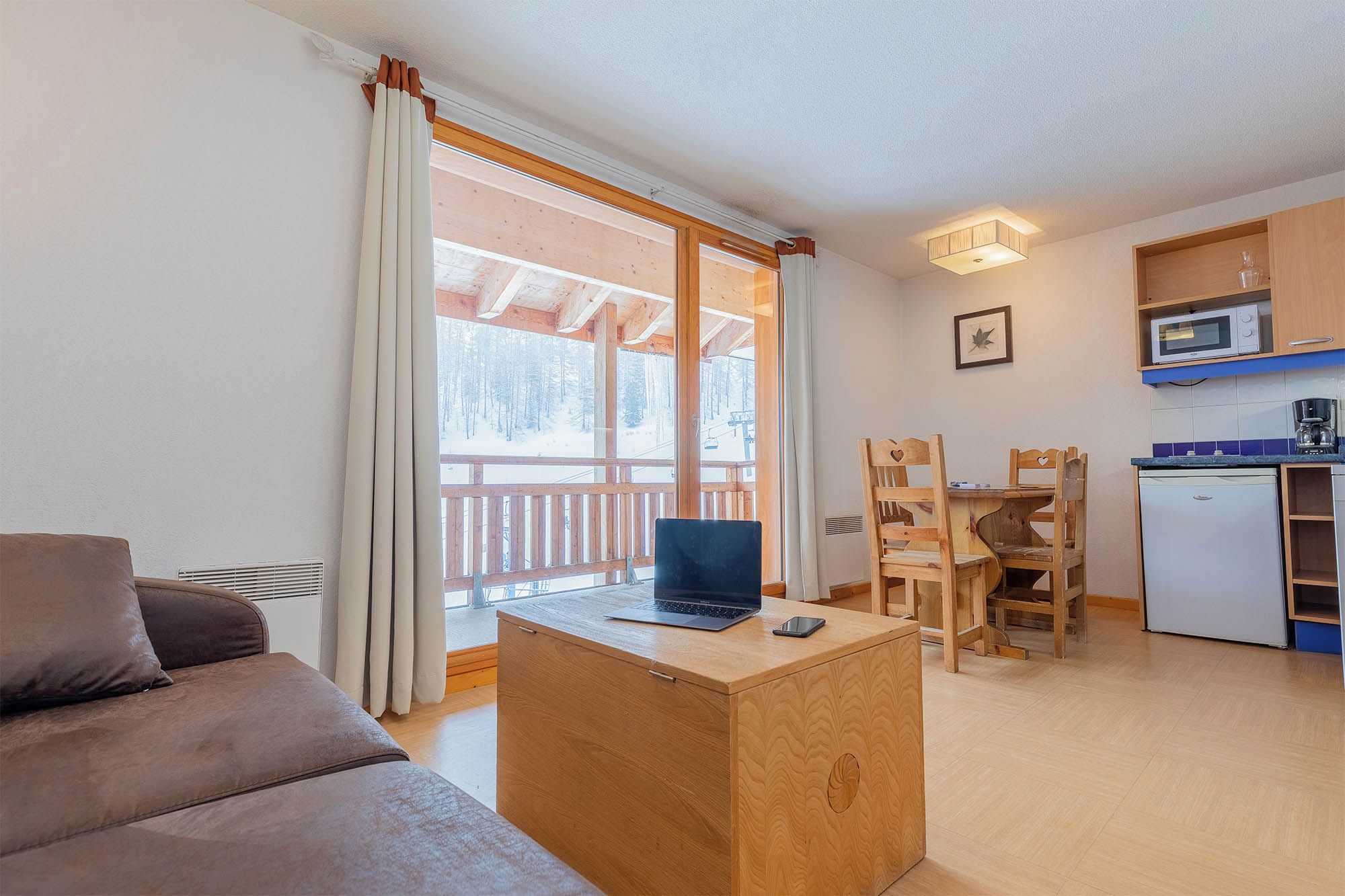 Ein gemütlicher Wohnbereich mit einer Couch, einem Holztisch mit einem Laptop und einem Esstisch mit Stühlen. Im Hintergrund ist eine Küche mit einer Mikrowelle und einem Kühlschrank erkennbar.