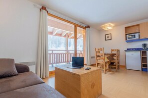 Een gezellig woongedeelte met een bank, een houten bureau met een laptop en een eettafel met stoelen. In de achtergrond is een keuken met een magnetron en een koelkast zichtbaar.