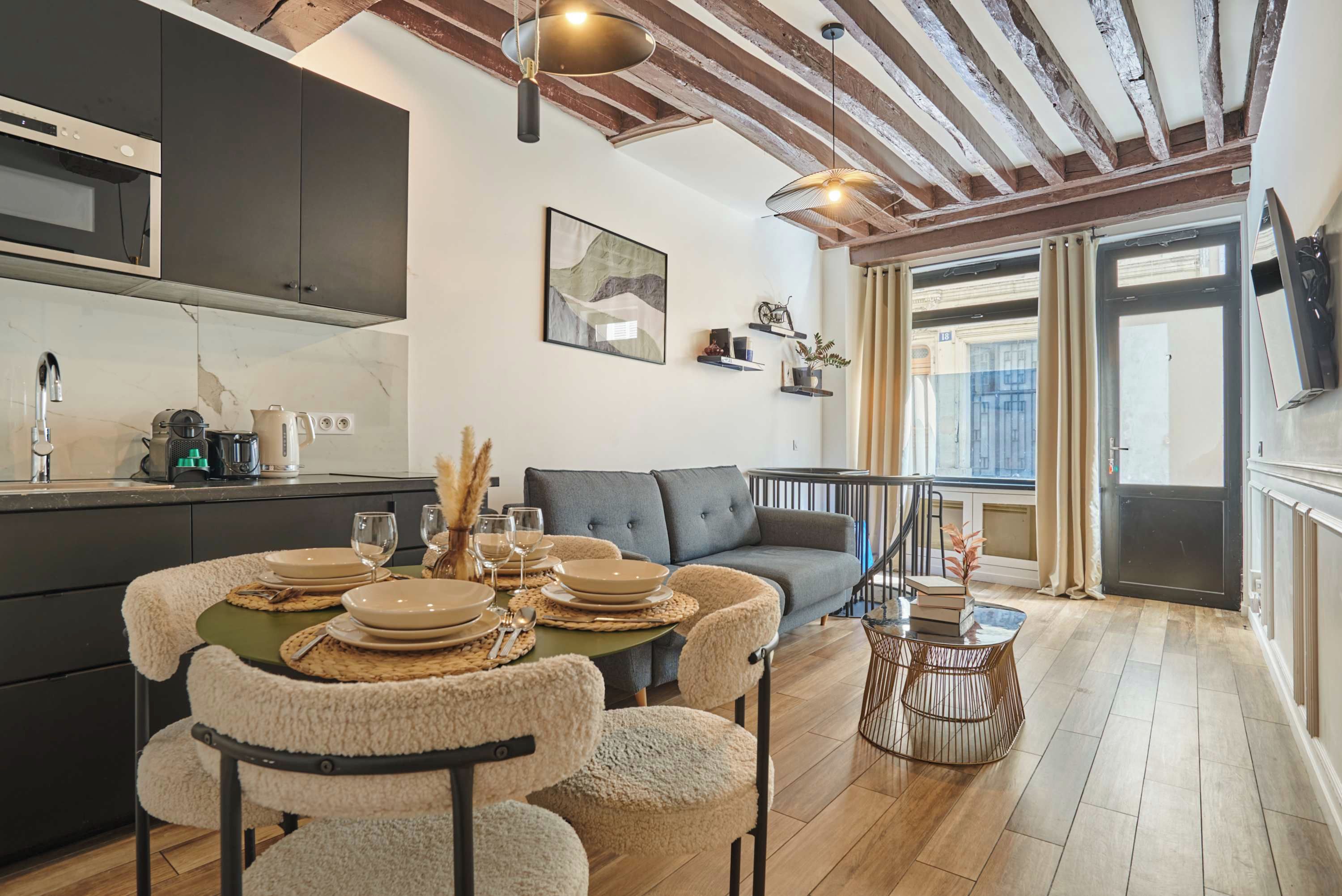 A modern kitchen with dark cabinets, a dining area with a round table set for four, and a living room with a sofa and a view of the city.