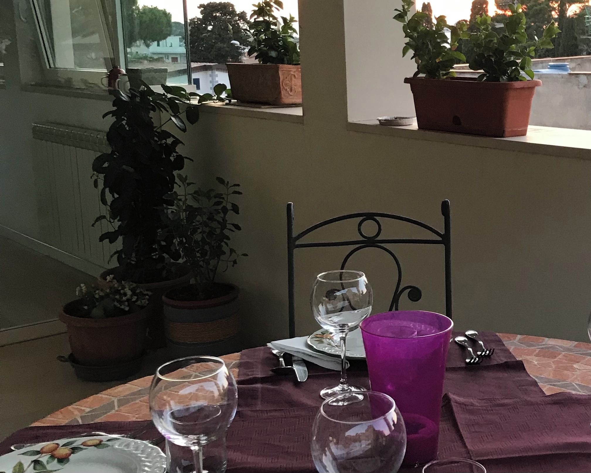 Ein Tisch mit einem Weinglas, einer violetten Tasse und einem Teller mit floralem Dekor. Auf dem Fensterbrett stehen Blumentöpfe.