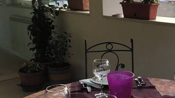 Ein Tisch mit einem Weinglas, einer violetten Tasse und einem Teller mit floralem Dekor. Auf dem Fensterbrett stehen Blumentöpfe.