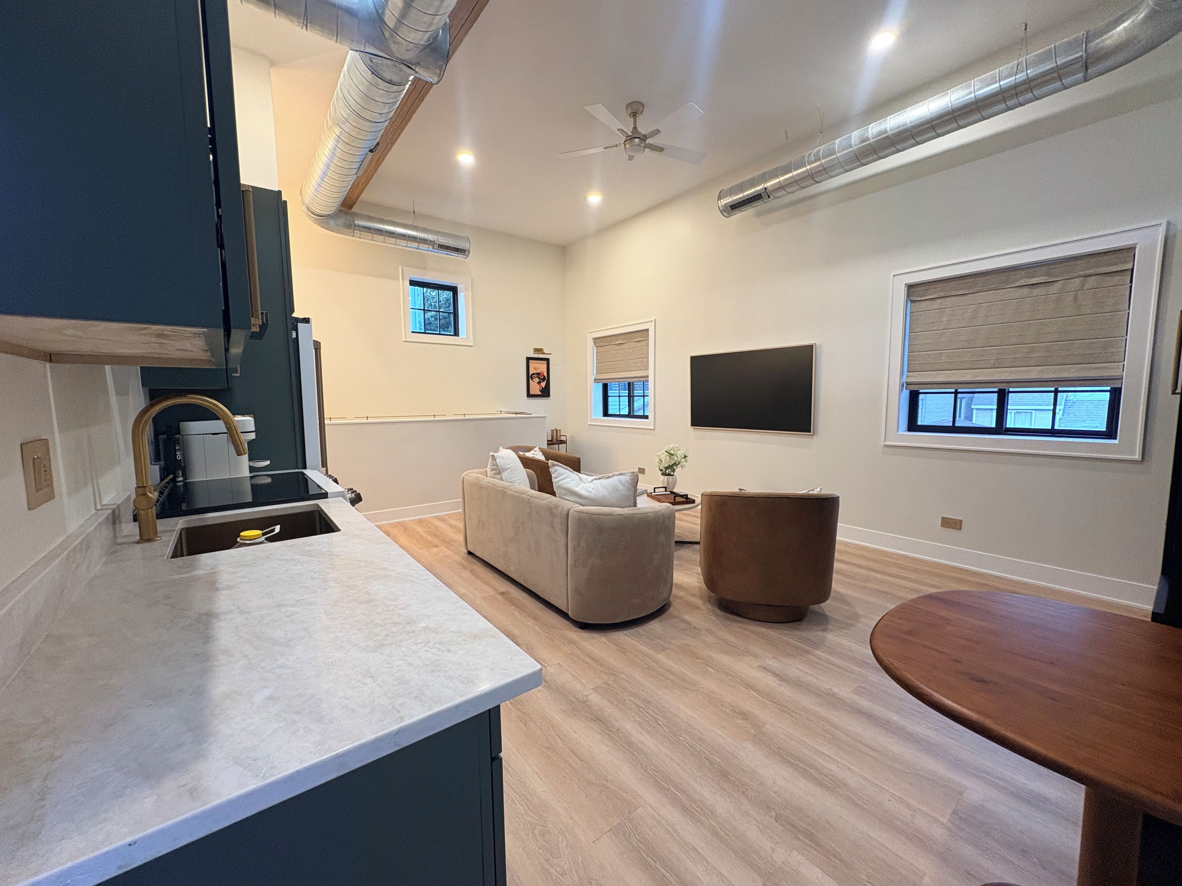 A modern kitchen with a built-in oven, a dining area with a wooden table, a sofa, and a TV mounted on the wall.