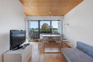Ein modernes Wohnzimmer mit Flachbildfernseher, Holzboden, einem Essbereich mit Tisch und Stühlen sowie einem großen Fenster mit Blick ins Freie.