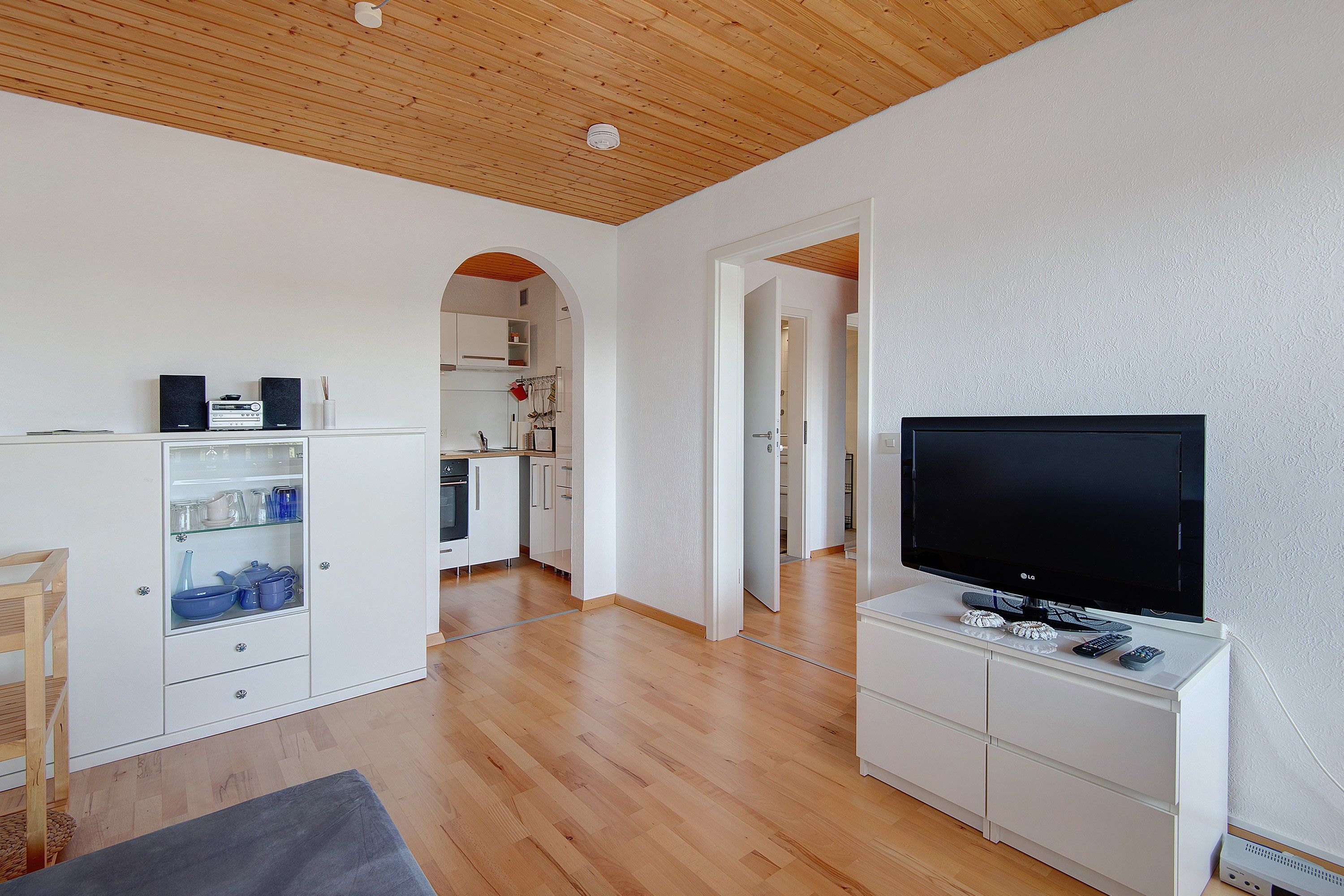 Un espace de vie moderne doté d’un téléviseur à écran plat, de planchers en bois, de meubles blancs et d’une porte en arc donnant sur un coin cuisine.