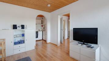 Un espace de vie moderne doté d’un téléviseur à écran plat, de planchers en bois, de meubles blancs et d’une porte en arc donnant sur un coin cuisine.