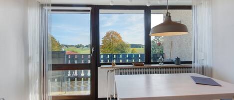 A room with a large window, a table, a bench, and a hanging light fixture.