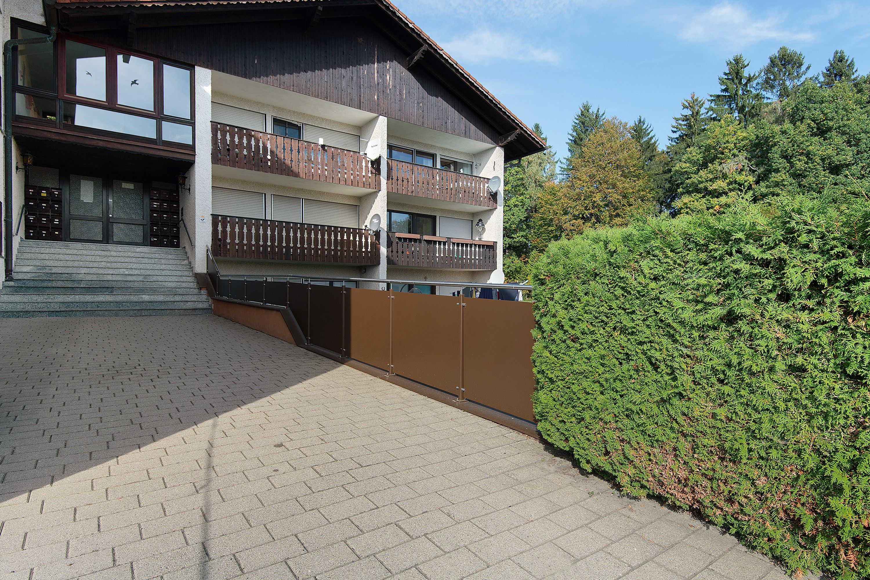 Ein mehrstöckiges Gebäude mit einem Balkon im zweiten Stock, einer Treppe, die zum Eingang führt, und einem gepflegten Heckenstreifen im Vordergrund.