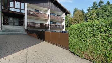Ein mehrstöckiges Gebäude mit einem Balkon im zweiten Stock, einer Treppe, die zum Eingang führt, und einem gepflegten Heckenstreifen im Vordergrund.