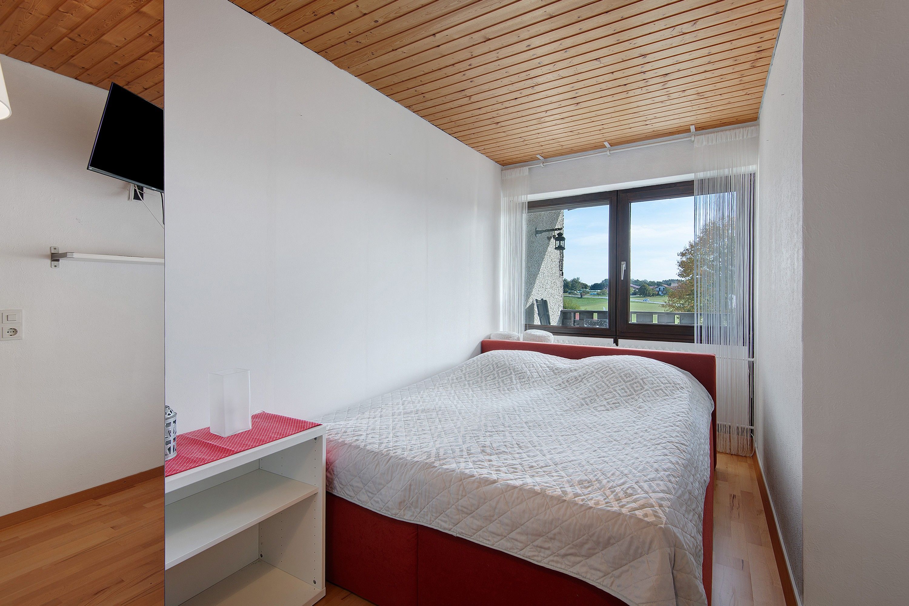 A bedroom with a bed, a window with curtains, a wooden ceiling, and a white wall.