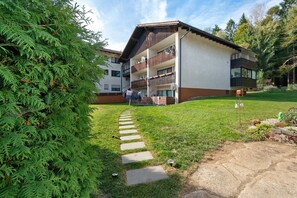 A multi-story building with a balcony, a grassy area with a stone pathway, and a dense green tree to the left. - Apartment 'Traum Im Bayrischen Wald' with Balcony and Wi-Fi (Wiesenfelden)