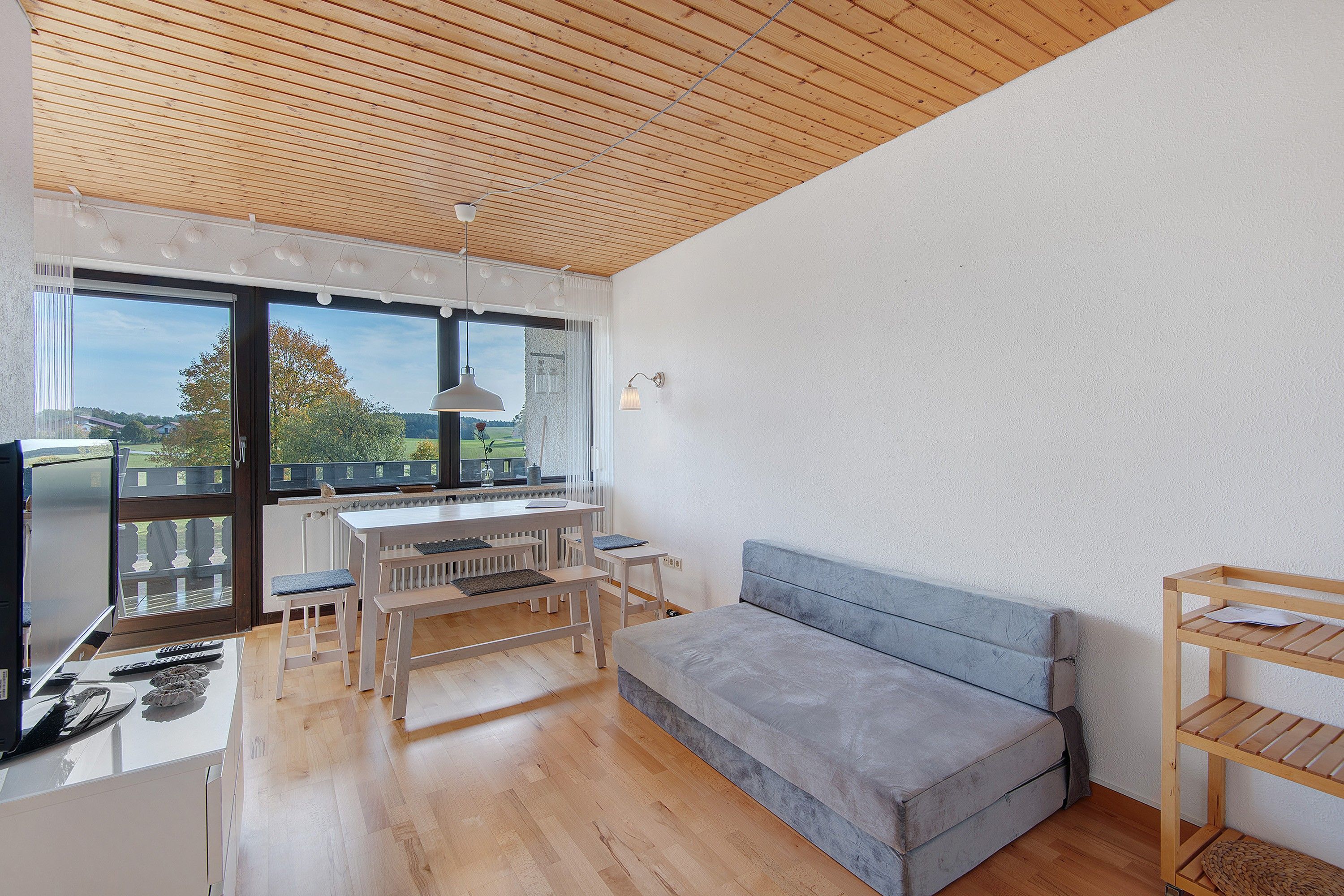 A modern living room with a wooden ceiling, a grey sofa, a dining table with benches, and a view of the outdoors through large windows.