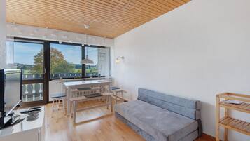 A modern living room with a wooden ceiling, a grey sofa, a dining table with benches, and a view of the outdoors through large windows.