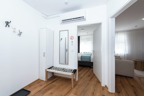 A modern hotel room with a wooden floor, a bench, a wardrobe, a bed with a blue blanket, and a bathroom with a mirror and a toilet. - Solentra - Popular City Location - Cozy 1BR - Free Garage (Vienna)