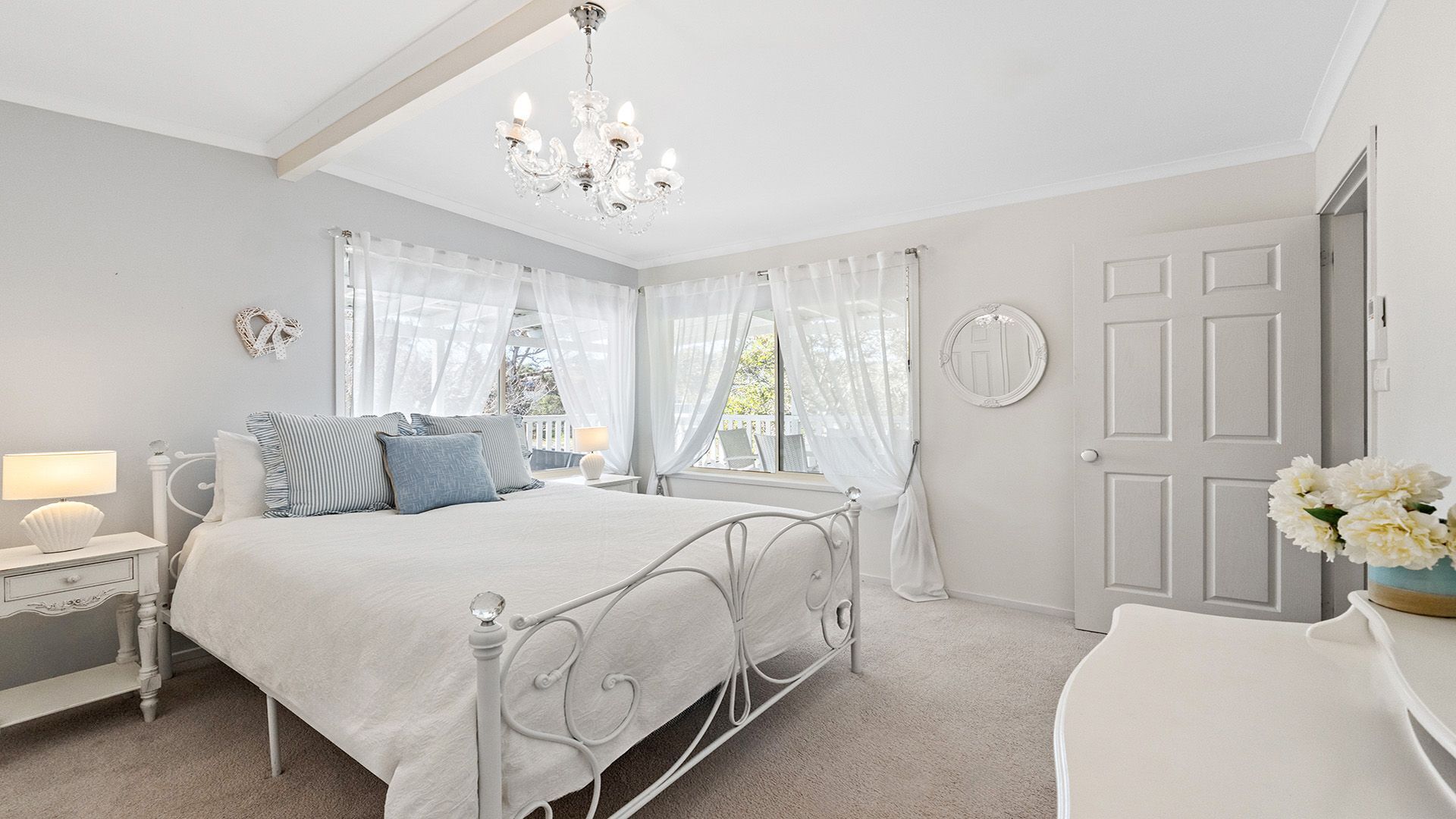 Une chambre avec un lit en métal blanc, une table de chevet avec une lampe, un lustre et une vue sur l’extérieur à travers des rideaux transparents.