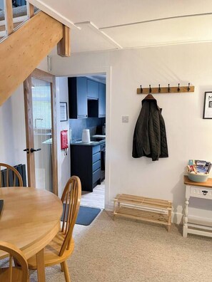 A room with a wooden dining table, chairs, a coat rack, a small bench, and a kitchenette in the background.