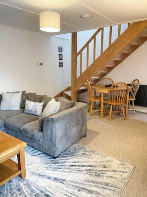 A living room with a sofa, a dining table with chairs, a staircase, and a hanging light fixture.