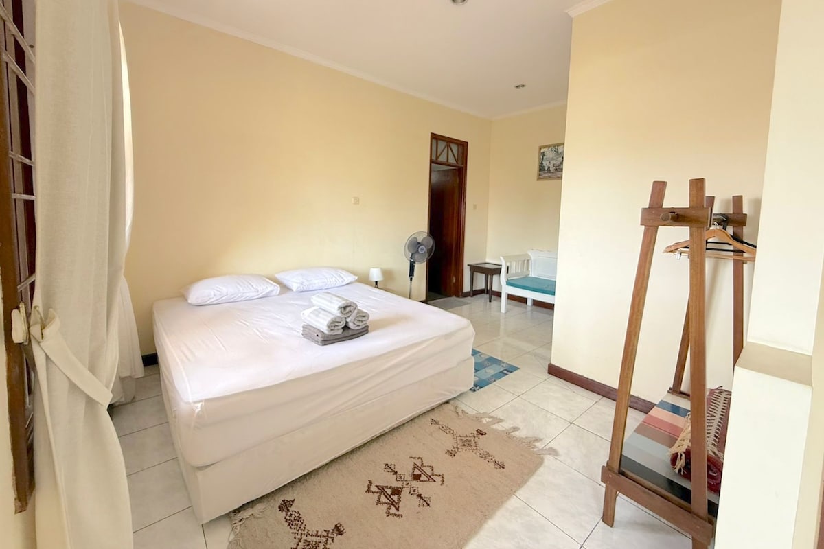 Una habitación de hotel con una cama, una mesita de noche, un ventilador, una silla, un perchero de madera y una puerta.