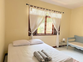 A room with a bed, a window with curtains, a bench, and a small table with a lamp.