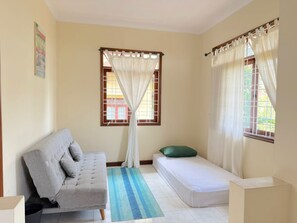 Una habitación con un sofá gris, una cama pequeña, una ventana con cortinas blancas y un tapete azul a rayas.