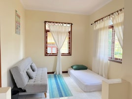 Una habitación con un sofá gris, una cama pequeña, una ventana con cortinas blancas y un tapete azul a rayas.
