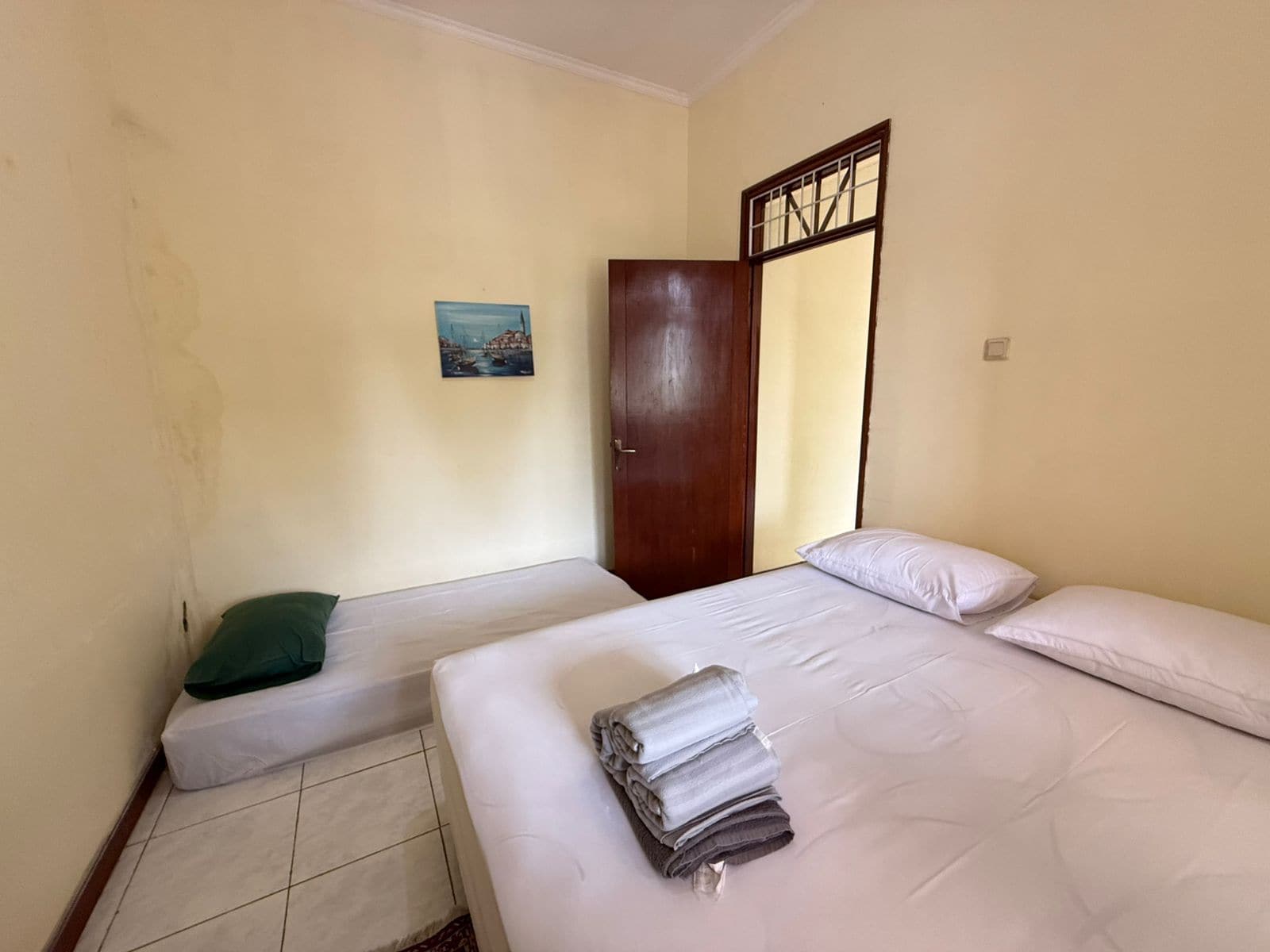 A room with two beds, a door, a painting on the wall, and a tiled floor.