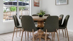 Een eetgedeelte met een ronde tafel, zes stoelen, een hangende rotan kroonluchter, een potplant en een ingelijst kunstwerk aan de muur.