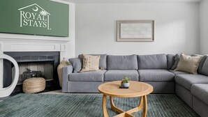 Een woonkamer met een grijze bank, een ronde houten salontafel, een open haard en een ingelijst schilderij aan de muur.