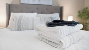 Ein ordentlich bezogenes Bett mit einem gefalteten weißen Handtuch und einem dunkelblauen Handtuch darauf, ein grauer gepolsterter Kopfteil und ein weißes Kissen mit geometrischem Muster.
